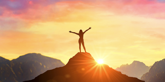 A woman is standing on top of a mountain with her arms outstretched at sunset.