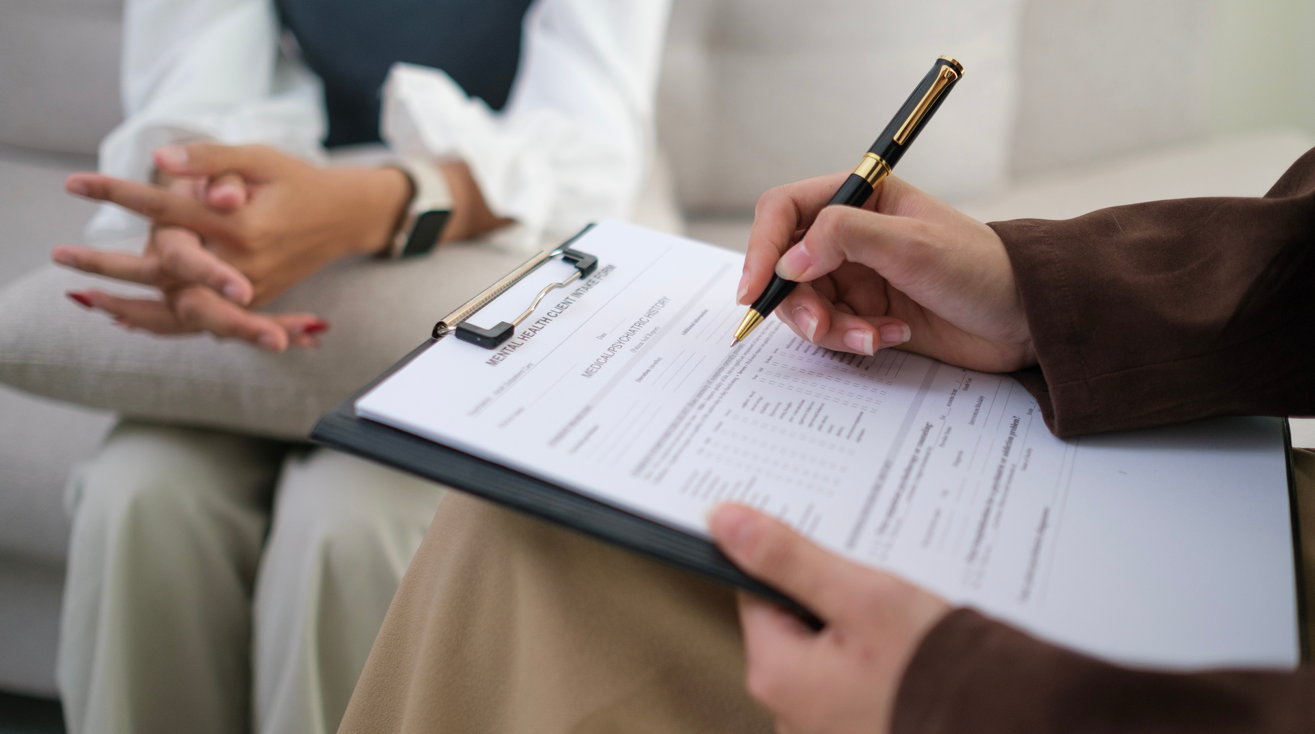 A woman is sitting on a couch while a man writes on a clipboard.