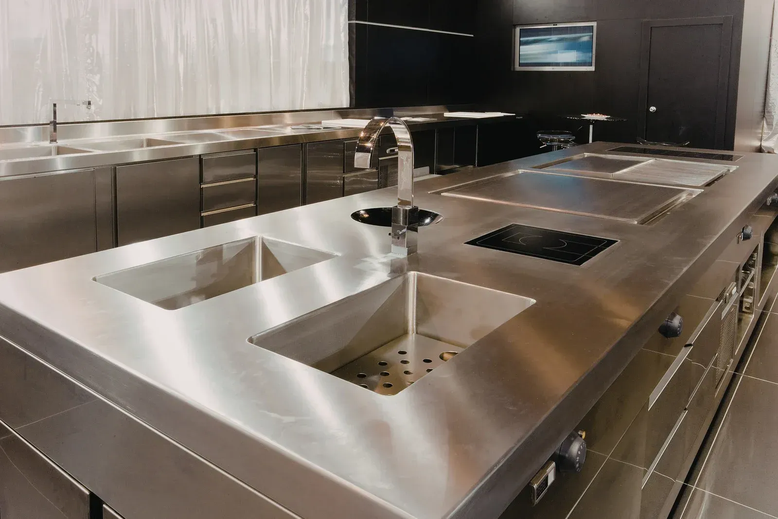A modern, professional kitchen featuring a large stainless-steel island with dual sinks, a faucet, and built-in stovetops.