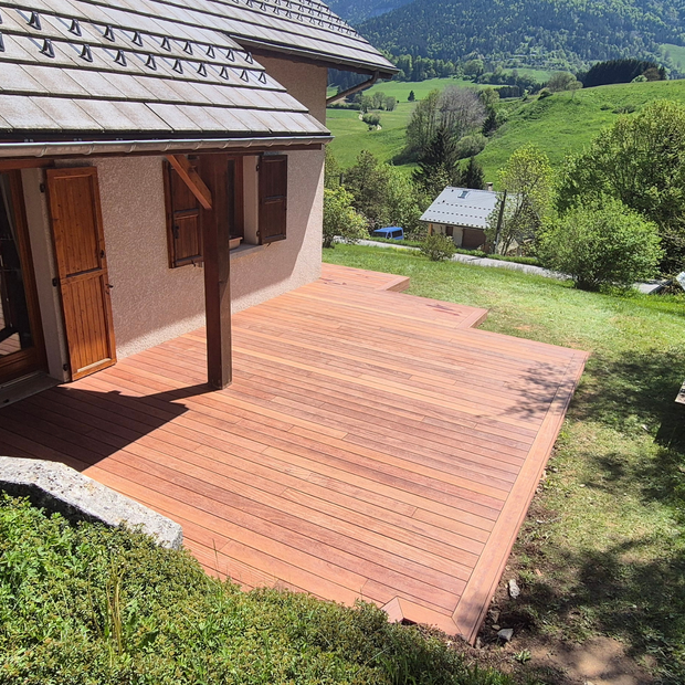 Terrasse en bois devant une maison avec un paysage montagneux et herbeux en arrière-plan.