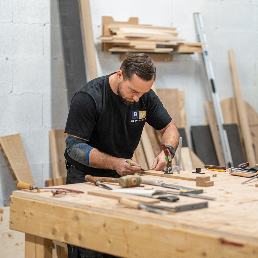 Dans un atelier, un homme marque du bois sur un établi entouré d'outils et de planches.