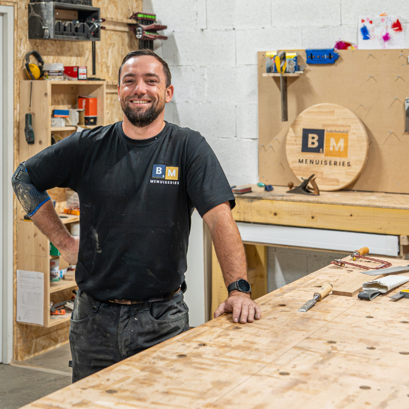 Un homme dans un atelier portant le logo « BM Menuiseries », appuyé sur un établi, souriant.