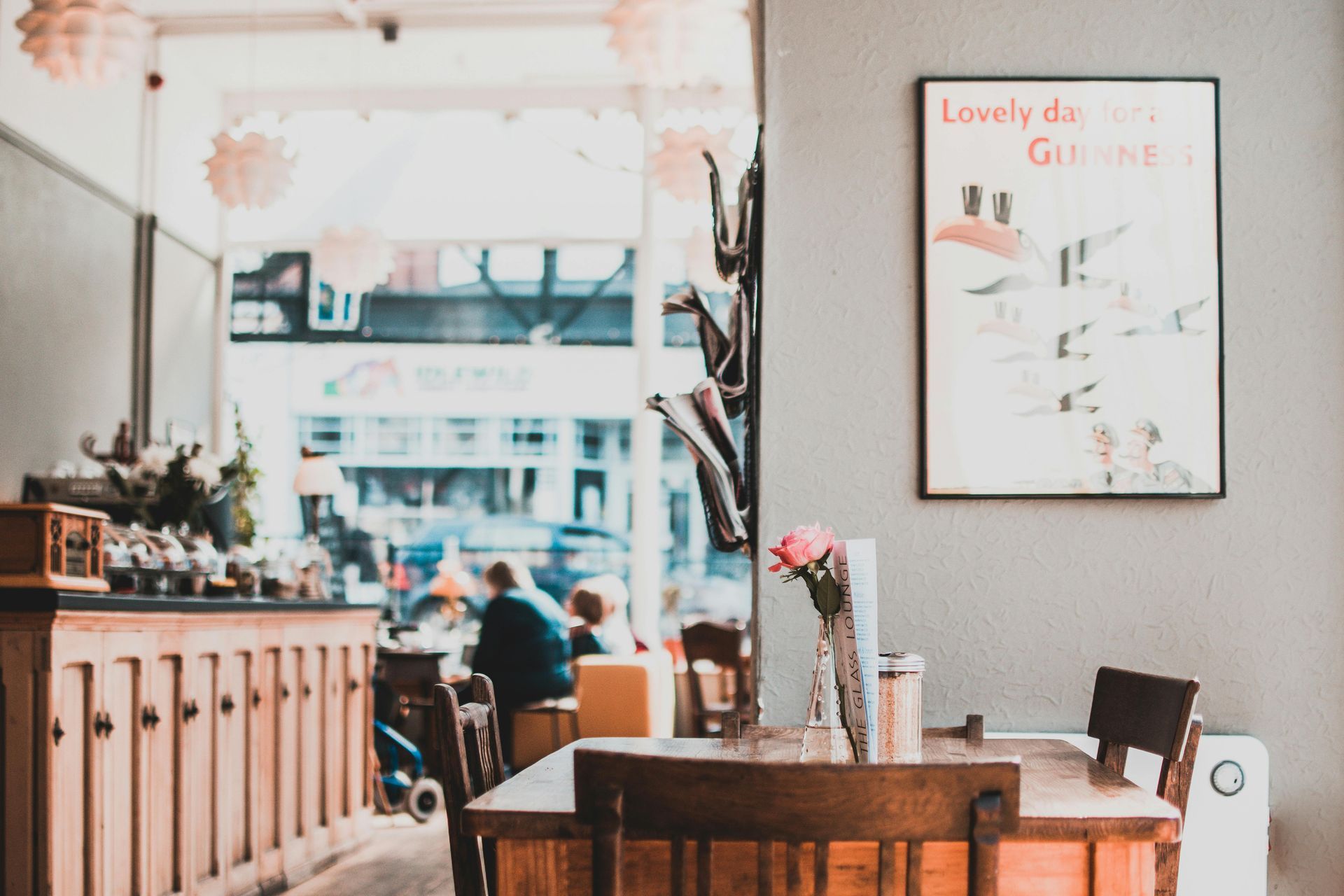 Interior cafe scene: table, chairs, framed artwork, flowers, wooden counter, window view of people outside.