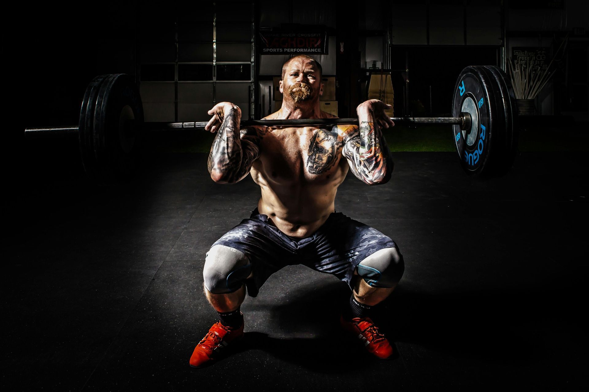 Man with tattoos squats while holding a barbell overhead in a dark gym.
