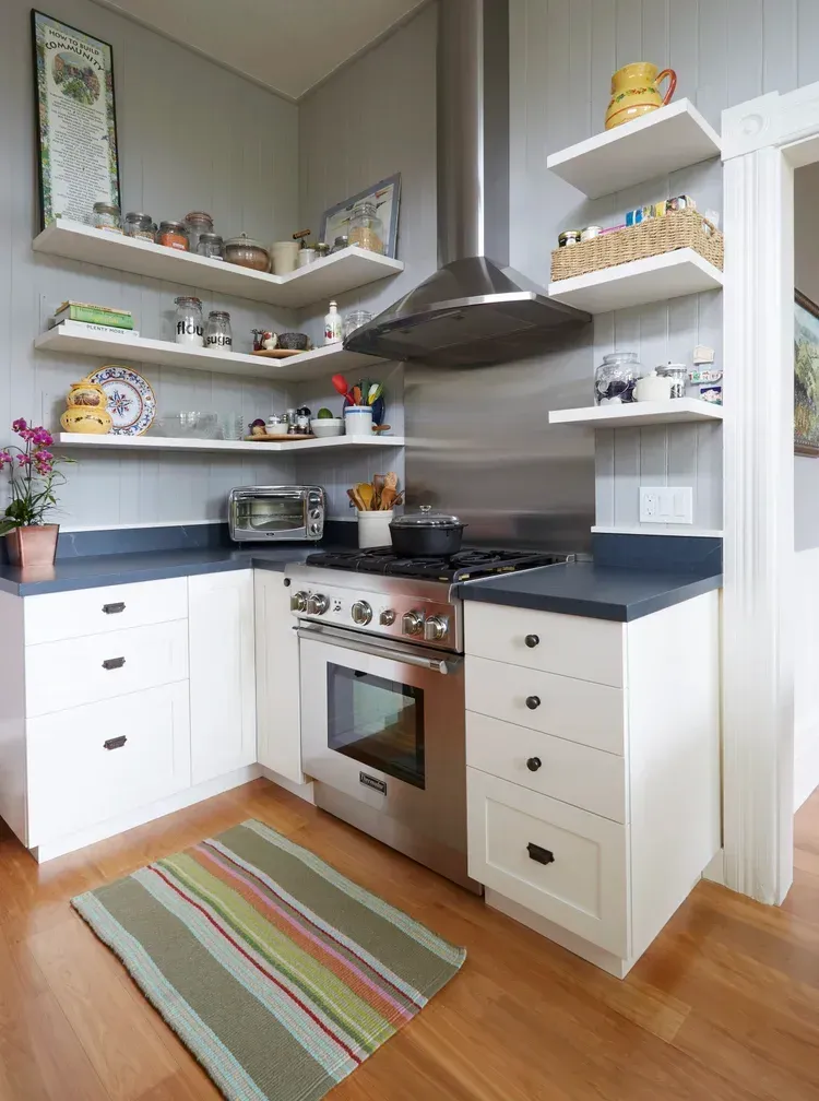 A small, bright kitchen with white cabinets, stainless steel appliances, and floating shelves filled with kitchenware.