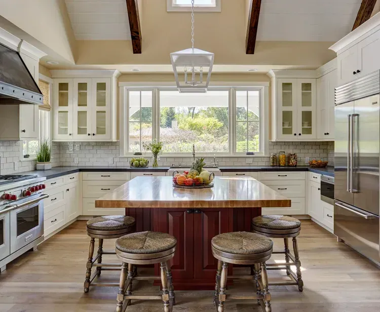 Kitchen with white cabinets, wood island with stools, and large windows.