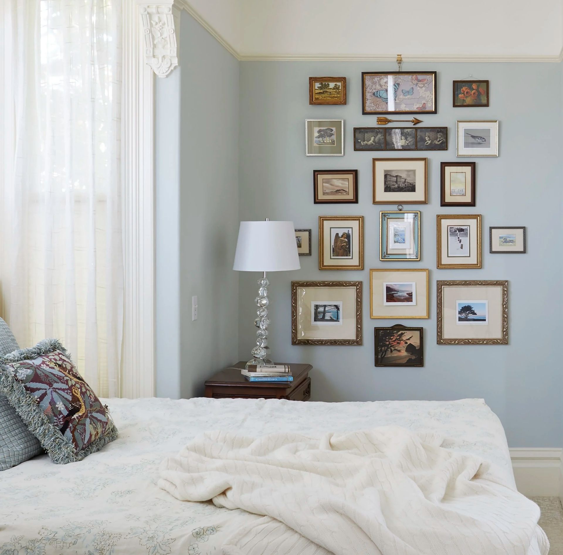 Bedroom corner with a gallery wall above the bed. Blue walls, a white bedspread, and a crystal lamp.