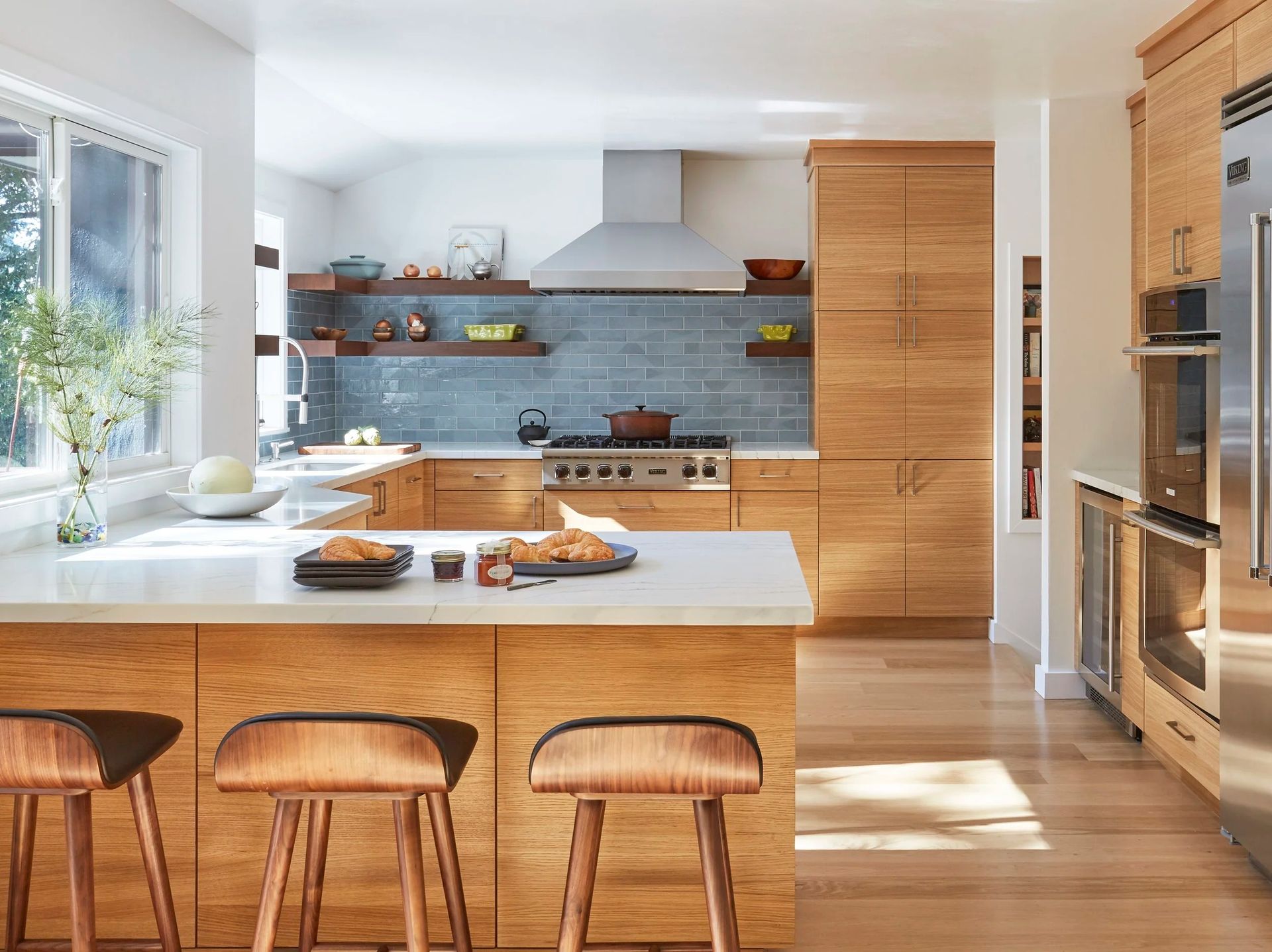 Modern kitchen with light wood cabinets, blue tile backsplash, stainless steel appliances, and a breakfast bar with stools.