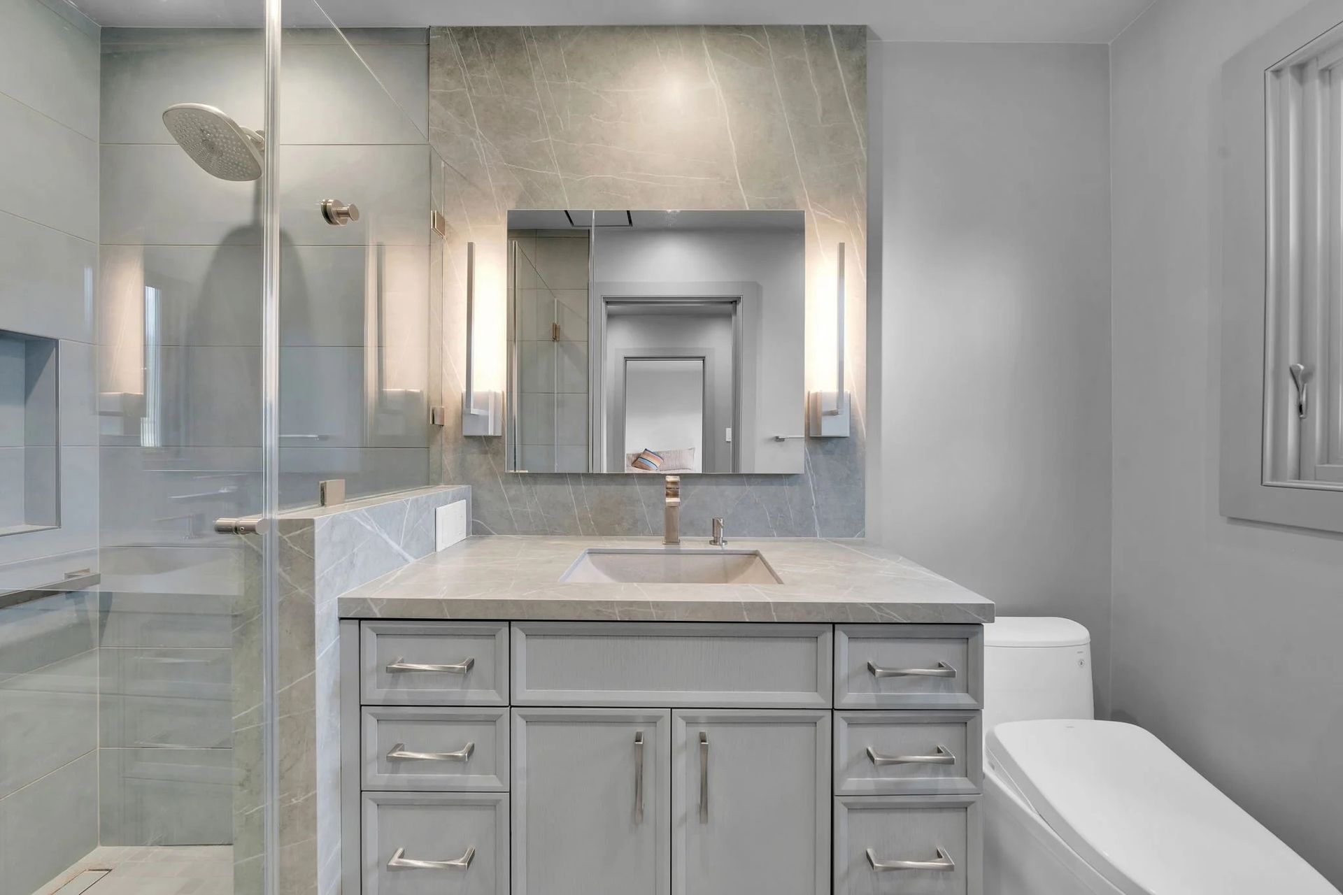 Modern gray bathroom with shower, vanity, toilet. Marble wall, light fixtures, glass door.
