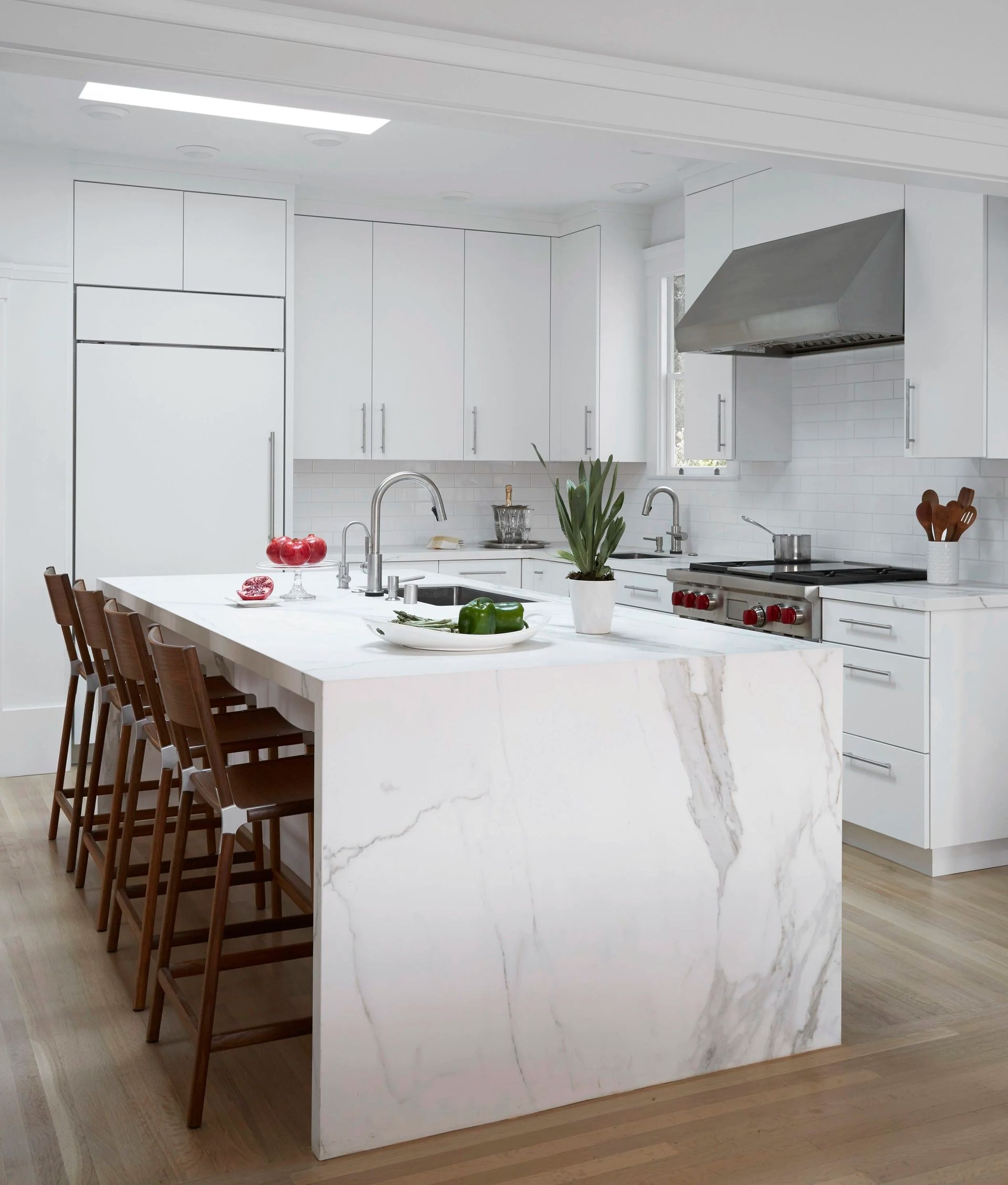 Modern white kitchen with marble island, stools, stainless steel appliances, and wood floors.