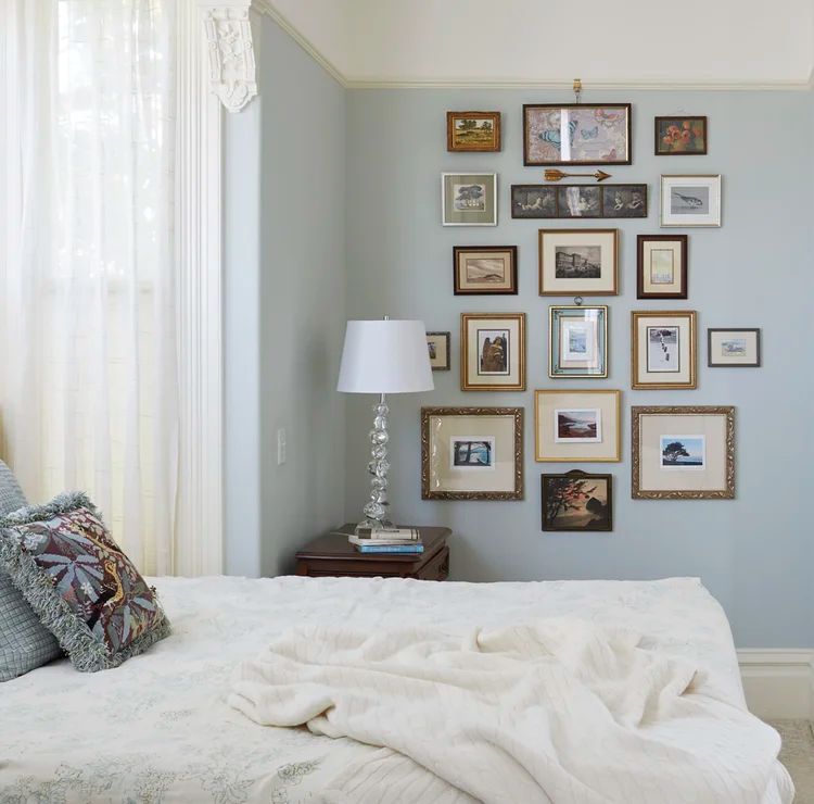 Bedroom with light blue wall, gallery wall of framed photos, bed with white bedding, and nightstand with lamp.