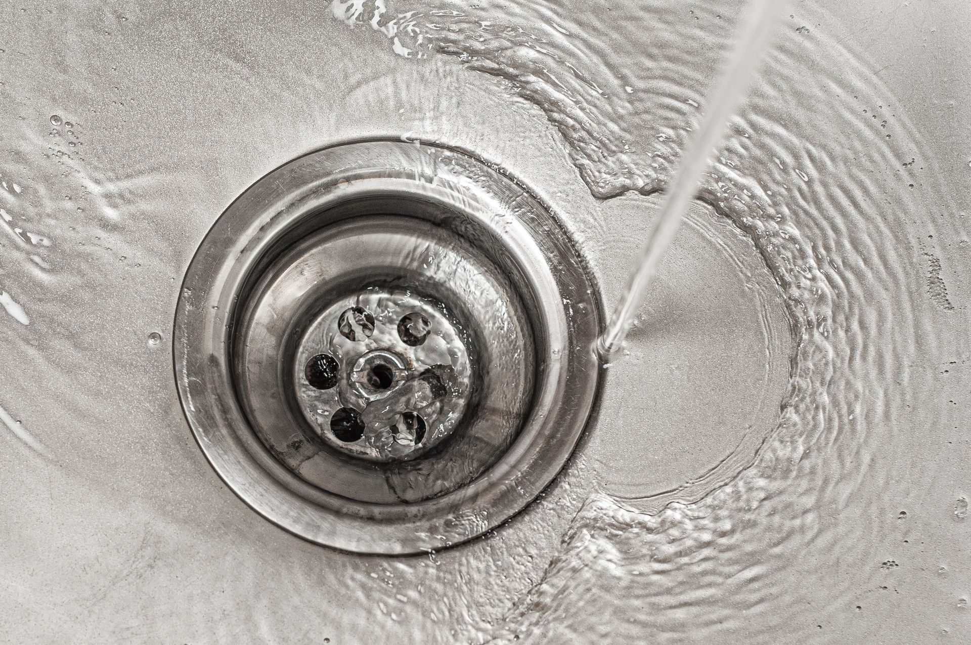 Water flows into a metal sink drain, creating ripples around the drain opening.