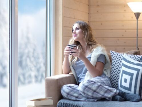 A woman is sitting on a couch holding a cup of coffee and looking out the window.