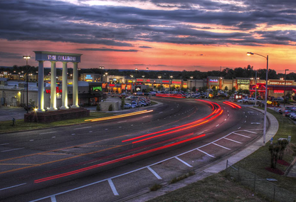 Sunset over a shopping center with blurred car lights on a curved road. Pillars mark the entrance.