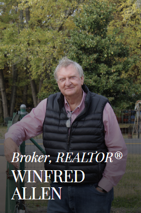 Broker Winifred Allen in black vest, arms resting on a fence, outdoors with foliage background.