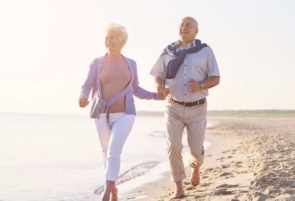An elderly couple is running on the beach holding hands.
