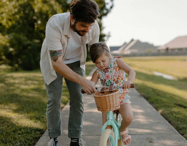 A man is helping a little girl ride a bike on a sidewalk.