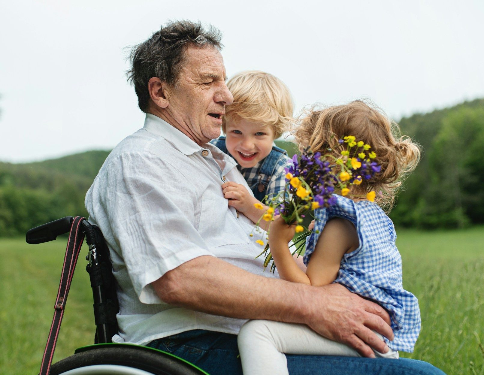 A man in a wheelchair is holding two children in a field.