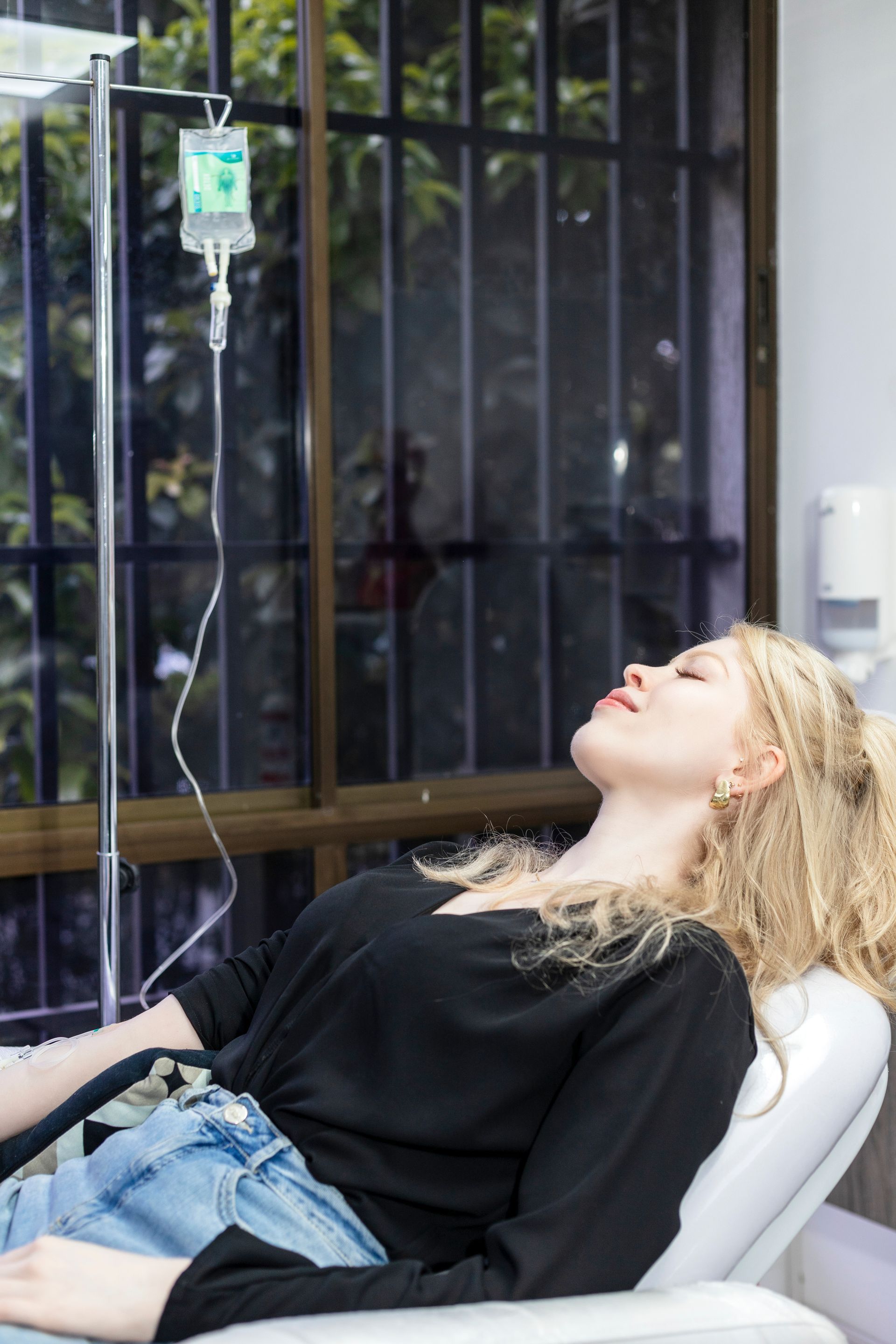 Mujer recibiendo goteo intravenoso en un entorno médico, con los ojos cerrados, cerca de una ventana.