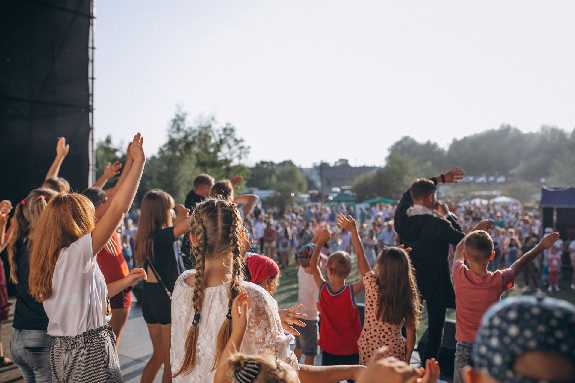 A diverse group of people at an outdoor concert with hands raised, facing a performer on a stage under a bright sky.