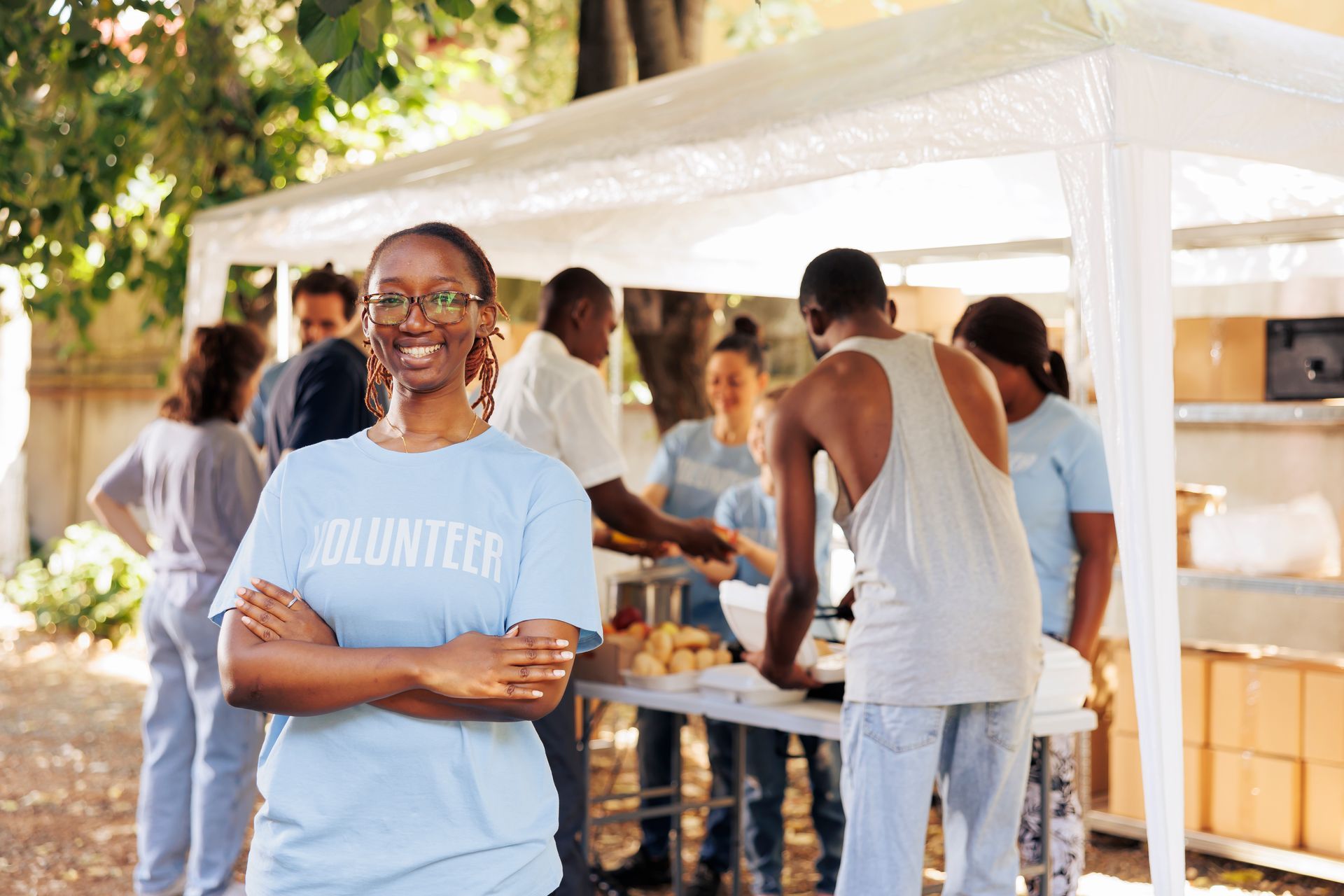 A smiling volunteer stands in the foreground with crossed arms; others prepare food under a white canopy in the background.