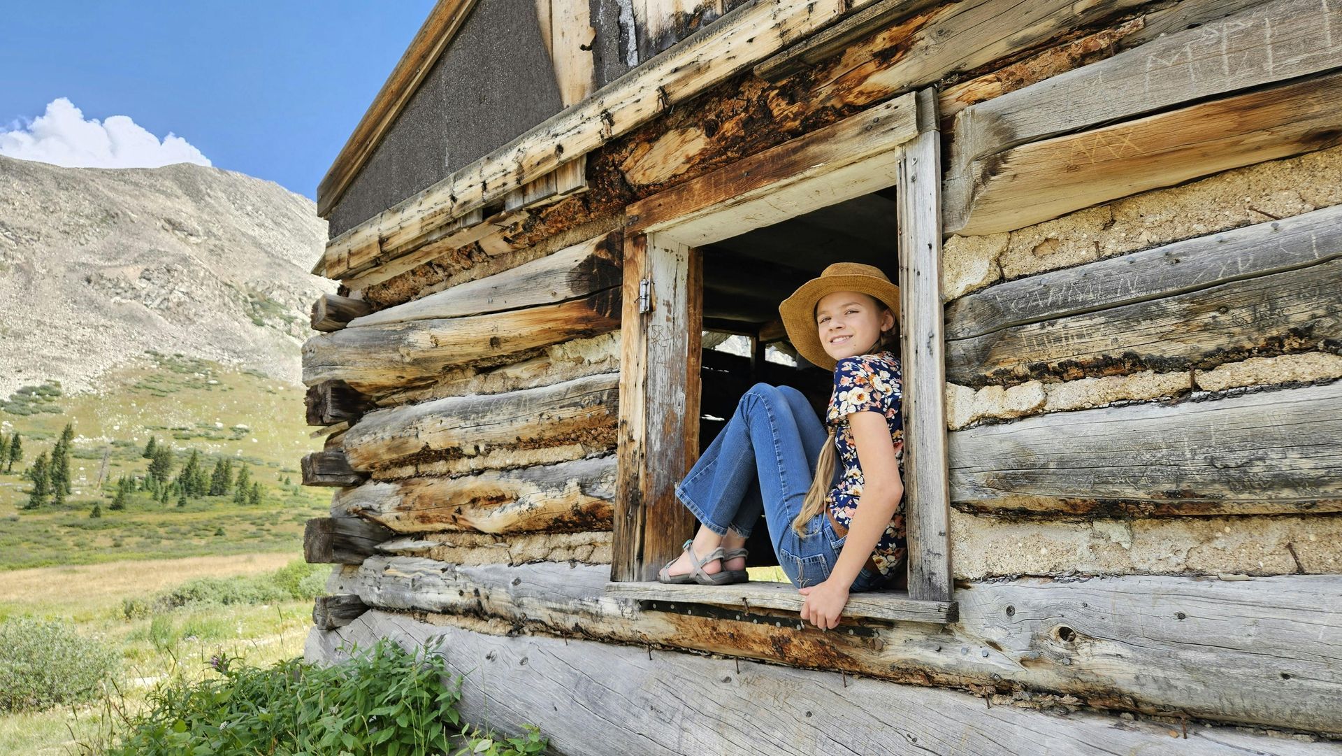 A person wearing a cowboy hat and jeans sits in the window frame of a rustic log cabin in a mountainous landscape.