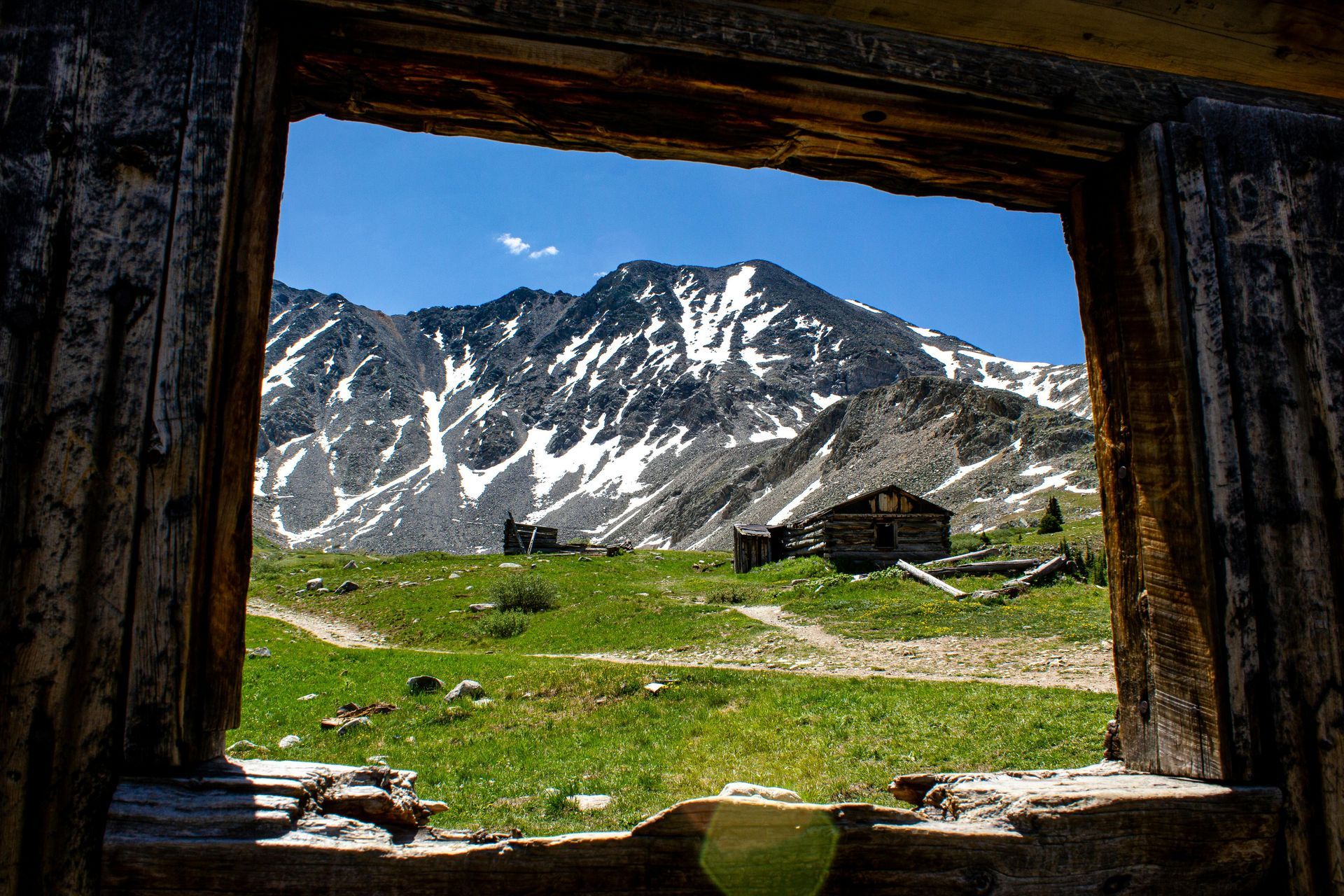 A view of snow-capped mountains and grassy hills seen through the weathered, wooden window frame of a ruined building.