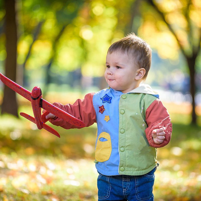 Young child in a green vest holding a red toy airplane in a sunlit park