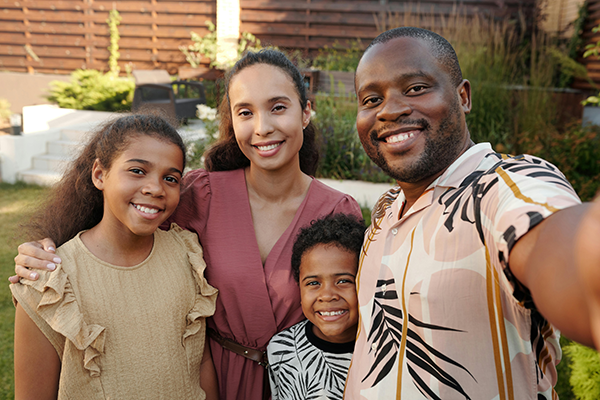 Four smiling people taking a selfie in a backyard, with a wooden fence and greenery behind them.
