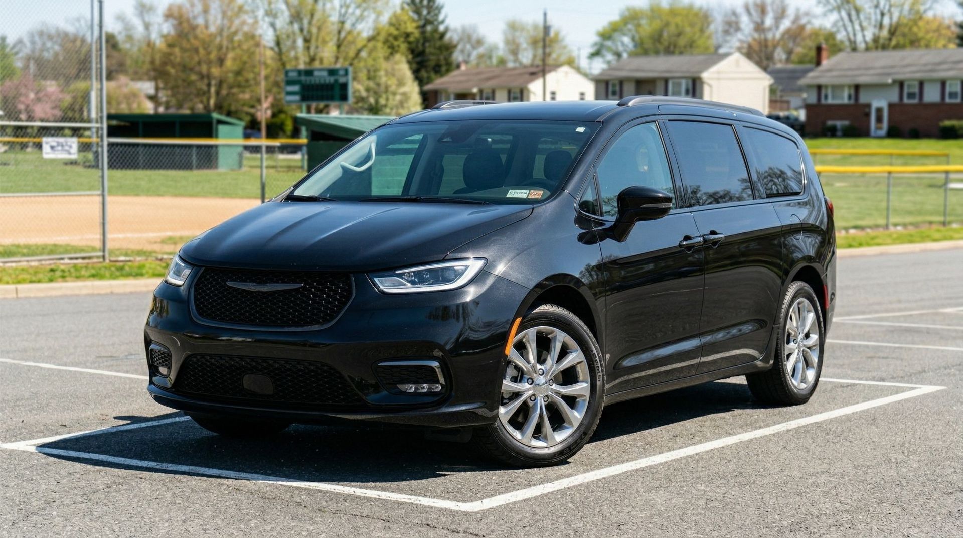 A black Chrysler Pacifica minivan parked in a paved lot near a sports field on a sunny day.