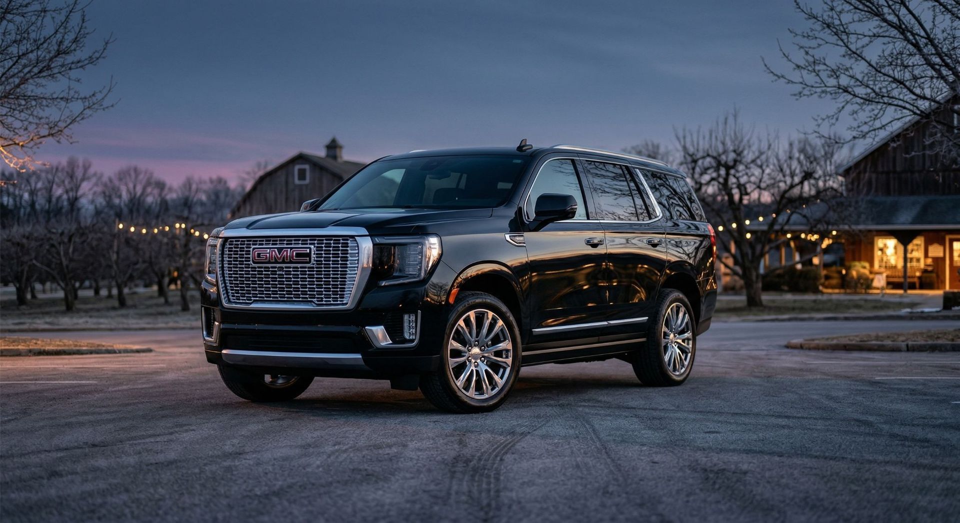 A black GMC Yukon SUV parked on a paved driveway in front of a rustic farm building at dusk.