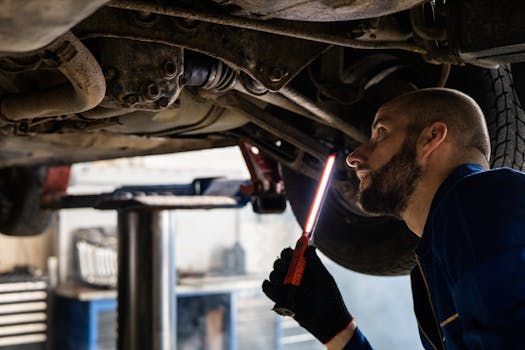 Mechanic in blue overalls inspecting under a car, using a flashlight.