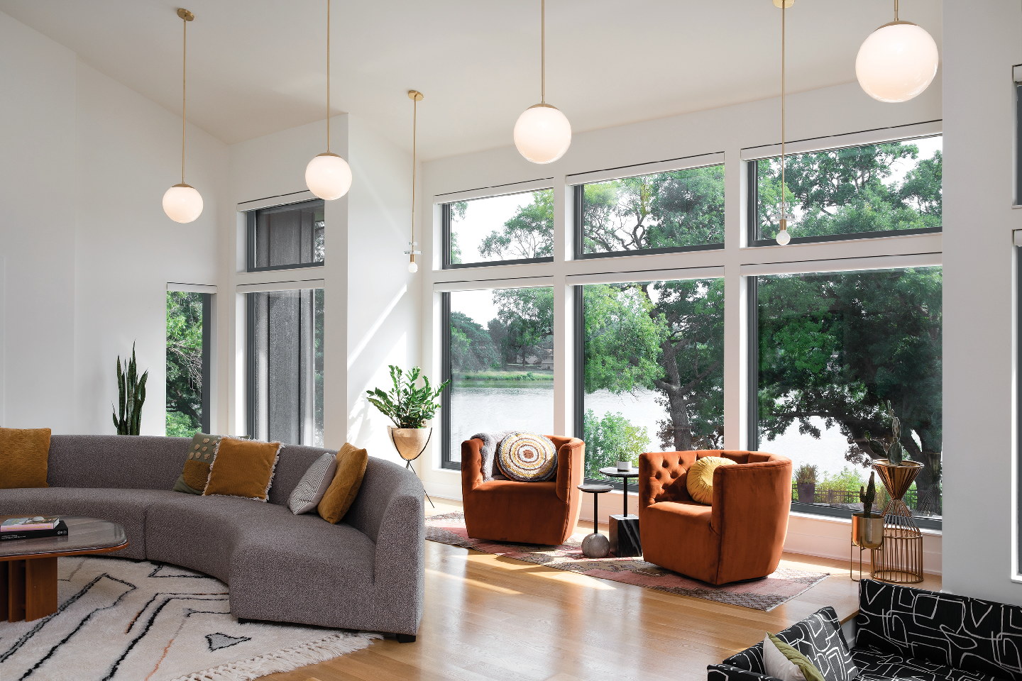 Modern living room with curved sofa, two orange armchairs, large windows, and globe pendant lights.