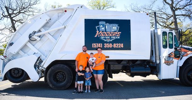 Garbage truck with three people standing in front, outdoors on a sunny day