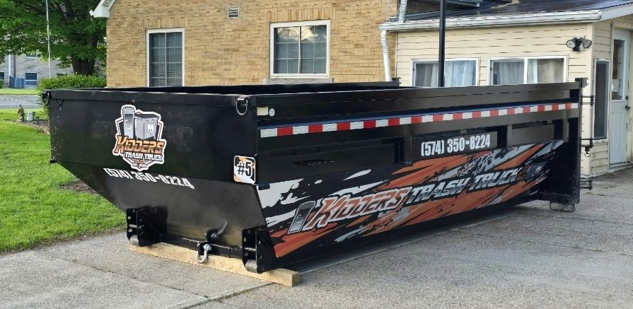 21.5 Yard Dumpster Black with orange-and-white business signage sitting beside a house and basketball hoop, with a red fire hydrant in front.