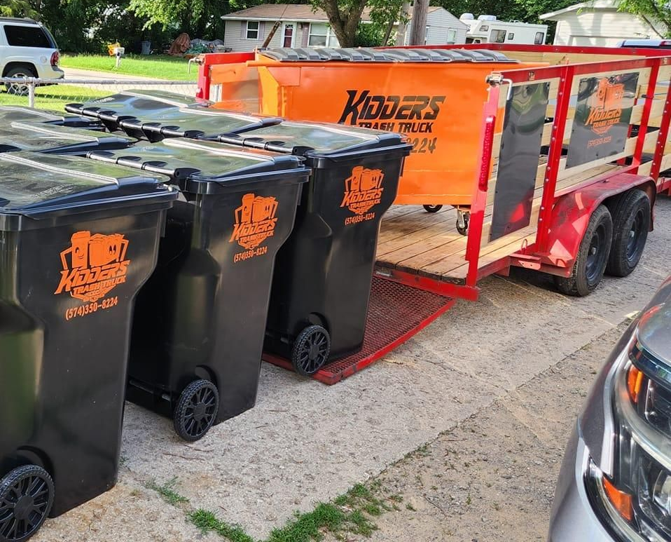 Black and orange trash bins beside a red Kidder's Trash Truck trailer on a residential street