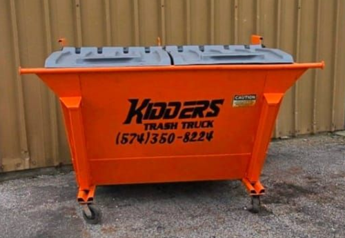 Orange Kidder's trash truck waste management company's 8 yard dumpster on wheels in front of a tan corrugated wall. 