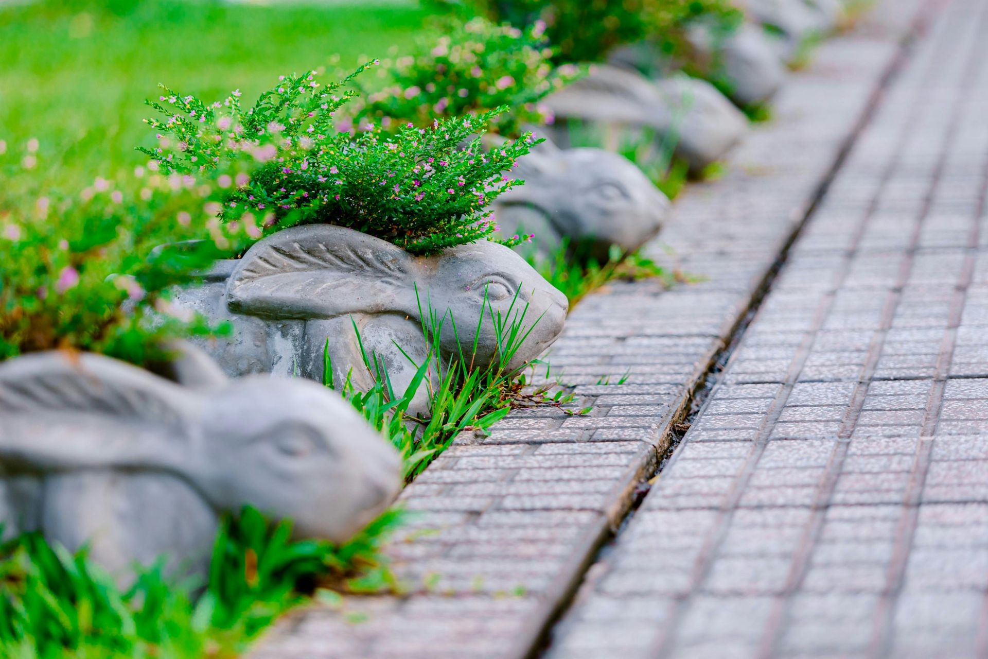 Row of concrete rabbit planters with greenery next to a brick walkway.