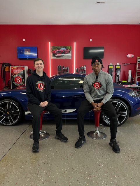 Two people sit in front of a blue sports car in a garage with red walls. Both wear matching logo hoodies.