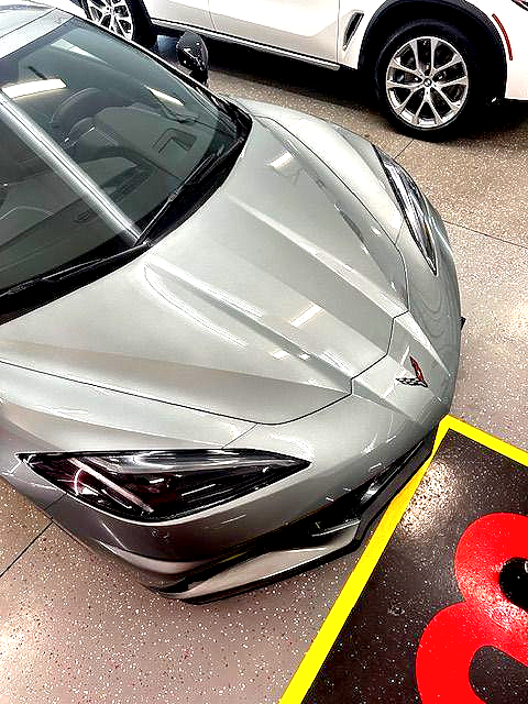 Silver sports car parked indoors, angled view of front. Yellow & black floor marker in view. White SUV in the background.