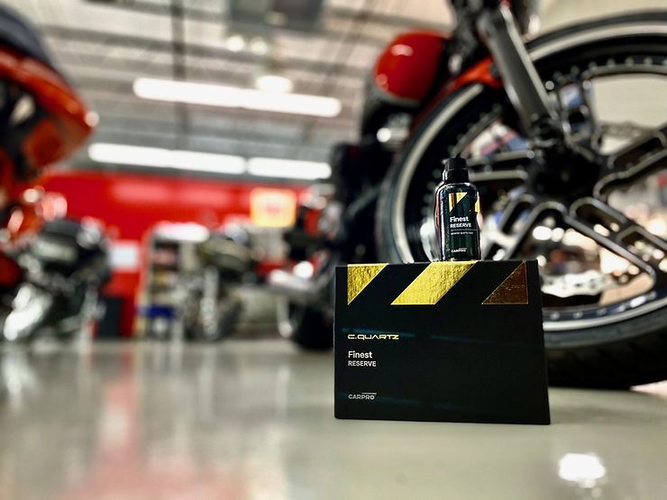 Black and gold product box and bottle on shiny floor with blurred motorcycle in background.