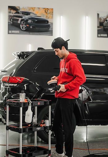 Man in red hoodie working on a black SUV in a detailing shop, holding a polisher.