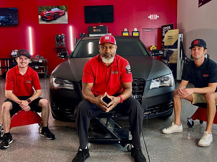 Three men in red shirts sit in a garage in front of a gray car. The garage has red walls.