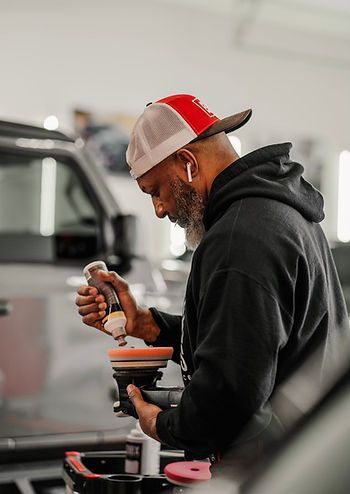 Man in black hoodie and red cap applies polish to car with polisher in shop.