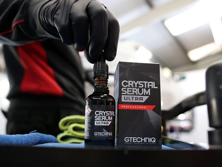 Person holding Gtechniq Crystal Serum Ultra bottle, next to its box; black gloves, auto detailing setting.