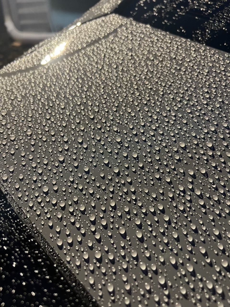 Black car surface covered in many water droplets, reflecting light.