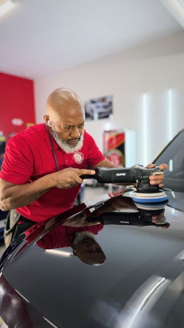 Man polishing a black car hood with an orbital buffer in a well-lit workshop.