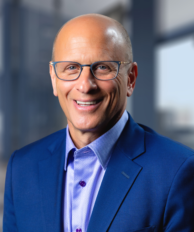 Harry Levant wearing glasses and a blue sweater is standing in front of a plant.