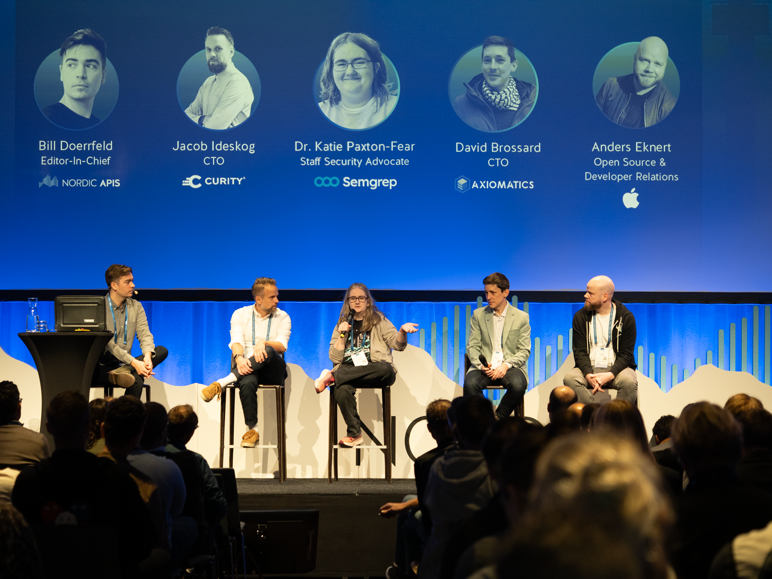 Panel discussion on stage with five speakers and audience in front of a blue screen with speaker names.