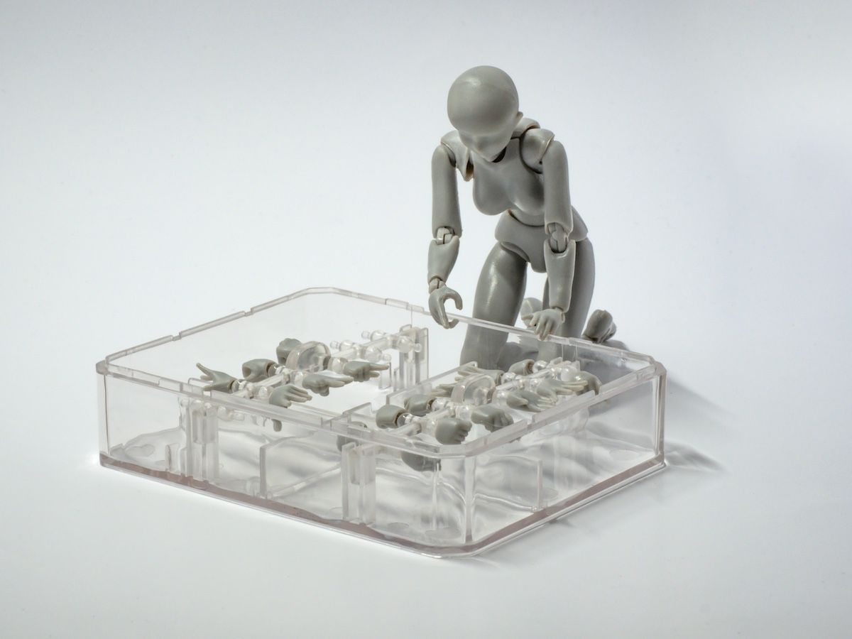 A grey articulated figure kneeling, arranging small white objects in a clear plastic container. White background.